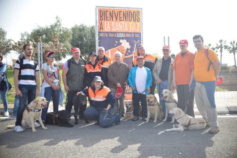 Grupo de personas con perros guía posa bajo el cartel «Bienvenidos a la Santa Faz».