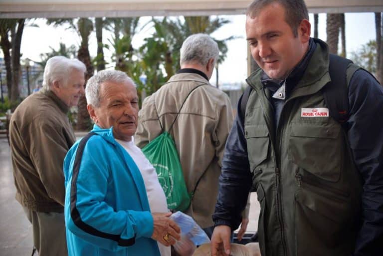 Varios hombres conversan en la calle; uno, con chaleco verde, sonríe en primer plano.