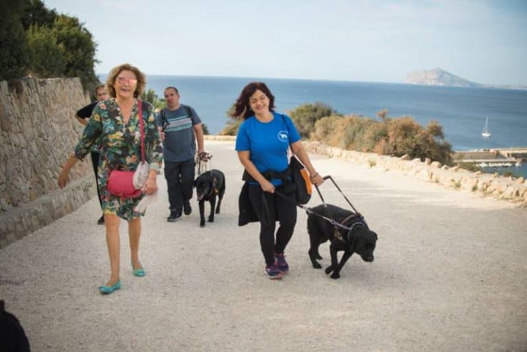 Tres personas caminan sonrientes con sus perros guía negros por un camino junto al mar, con un islote al fondo.