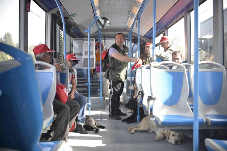 Interior de un autobús urbano: varias personas con gorra roja de pie y sentadas, y un perro guía labrador claro tumbado en el pasillo.