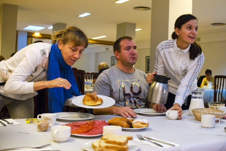 Desayuno en un comedor con mantel blanco: una mujer se sirve bollería, un hombre espera sentado y otra mujer sujeta una cafetera.