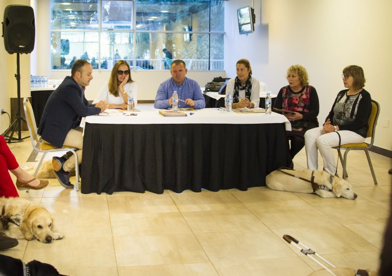 Mesa de ponentes con mantel negro en un salón; delante, dos perros guía claros tumbados en el suelo.