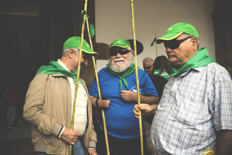 Tres hombres con gorra verde, pañuelo verde y cañas conversan a la sombra.