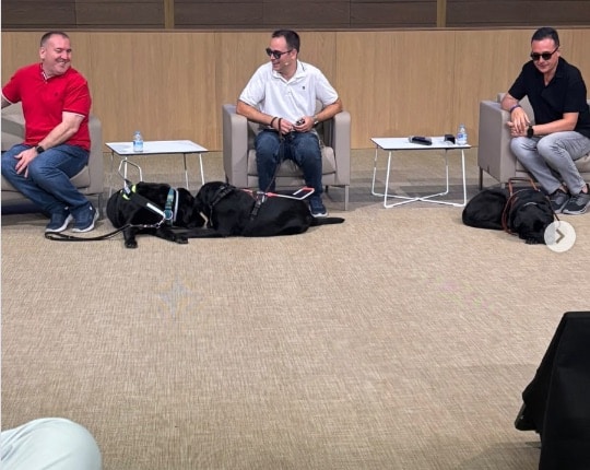 Tres personas usuarias de perro guía sentadas en un escenario del CEU, con sus perros tumbados a los pies.